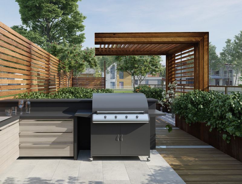 A 3D rendering of a modern outdoor kitchen on a wooden patio. It features a gas grill, dark countertops, a wooden pergola, and green plants.