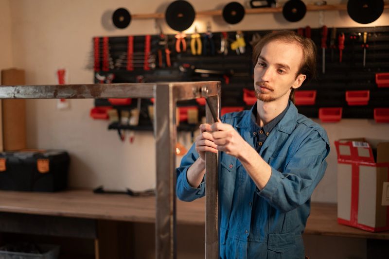 Male craftsman using wrench for metal table assembly in workshop with tools hanging on the wall. Professional handyman building steel furniture. Manual work and craft process.