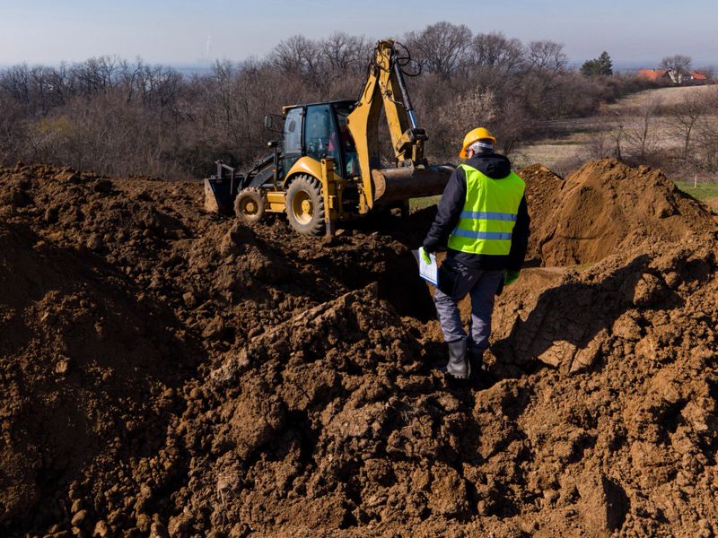 A construction engineer wearing protective gear stands on a muddy earthworks site and supervises a backhoe loader during excavation work.
