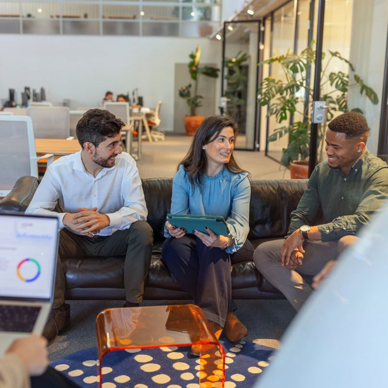 A diverse group of colleagues sits on a lounge sofa in a modern office, collaborating over a tablet and laptop, sharing ideas, relaxed conversation and friendly teamwork in an open-plan workspace.