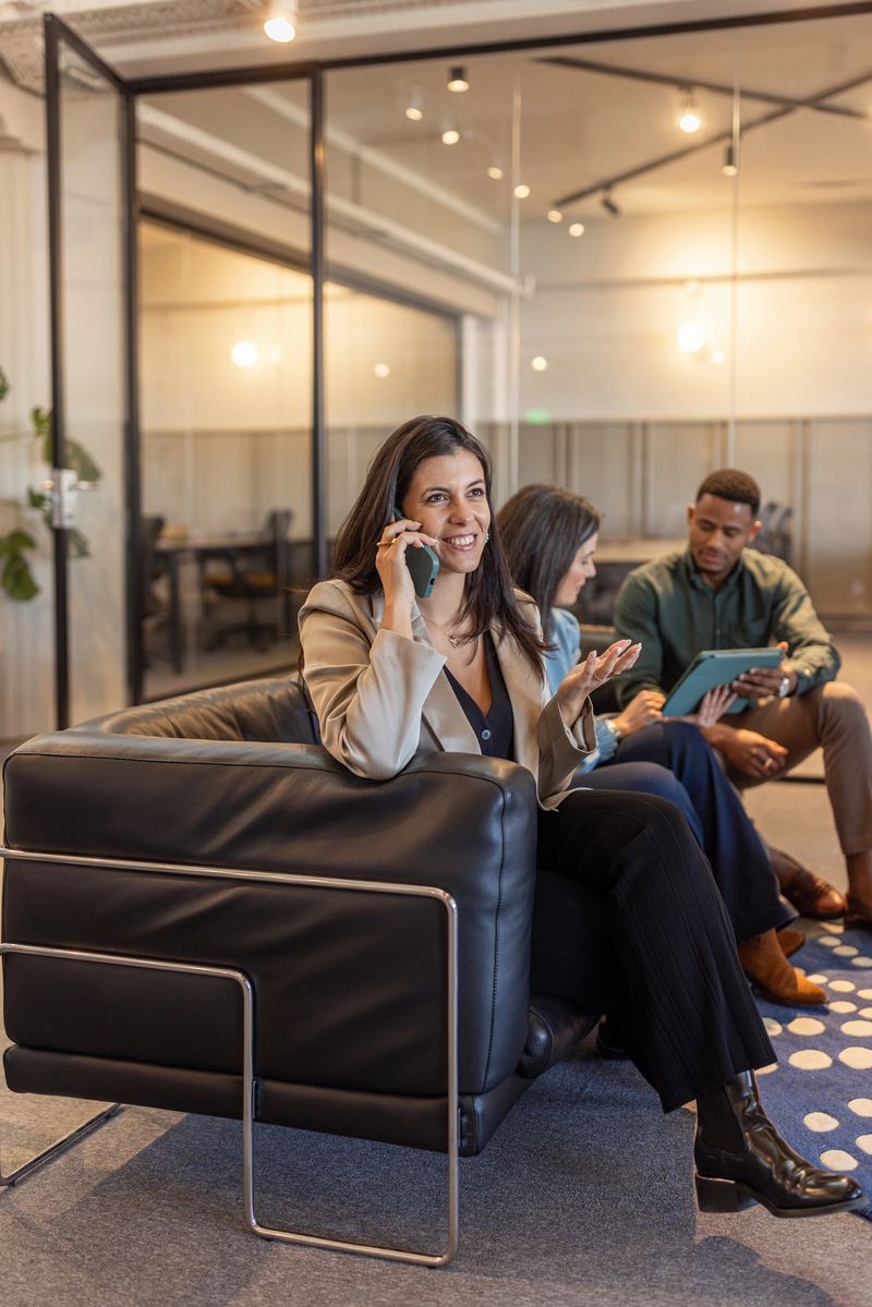 Smiling businesswoman talks on phone in a modern open office lounge while colleagues review a tablet nearby, conveying teamwork, collaboration, and a relaxed professional atmosphere in a corporate setting.
