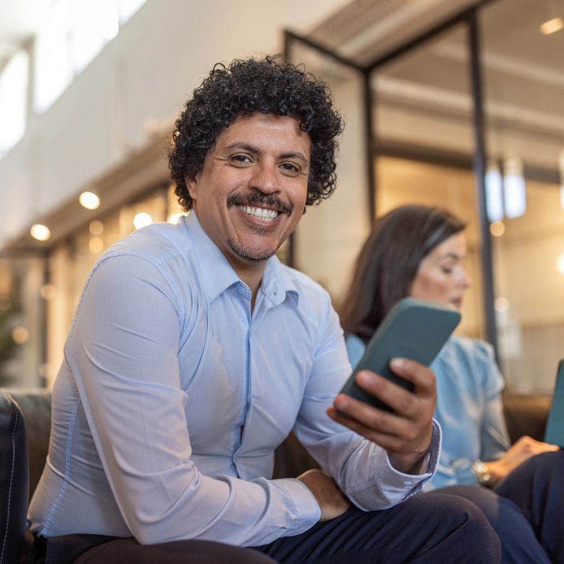 Smiling businessman sitting in a modern office waiting area, holding a smartphone while a coworker works in the background, conveying confidence, approachability, and professional collaboration.
