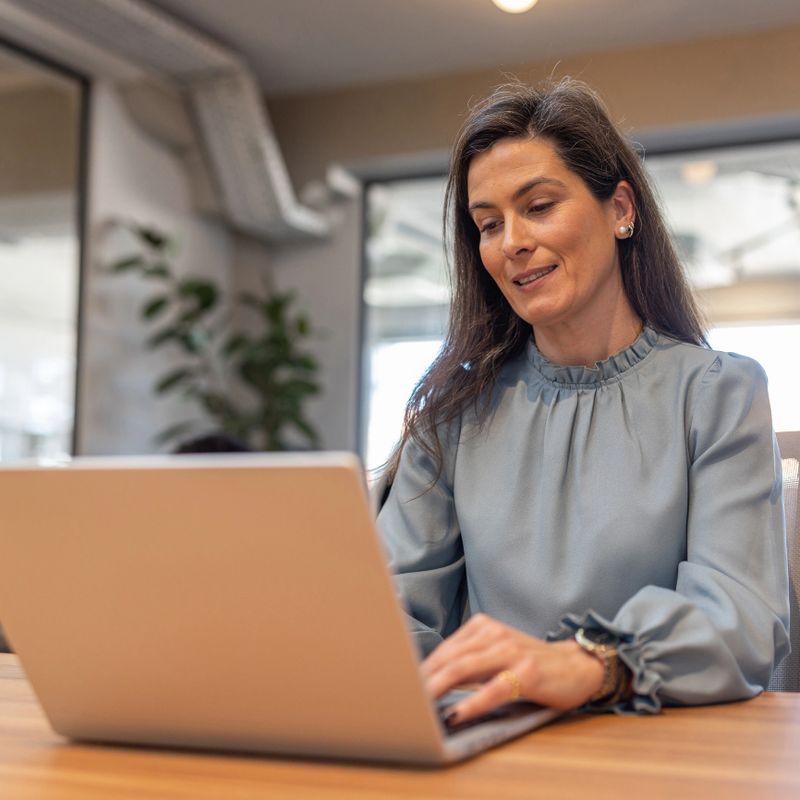 Professional woman typing on a laptop in a bright modern office, smiling and focused while working remotely; conveys productivity, confidence, hybrid work and positive business communication.