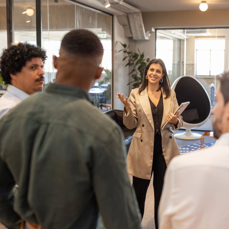 Confident female team leader presents ideas to colleagues in a modern open-plan office, holding a tablet and engaging the team in a collaborative meeting focused on strategy, communication and workplace culture.