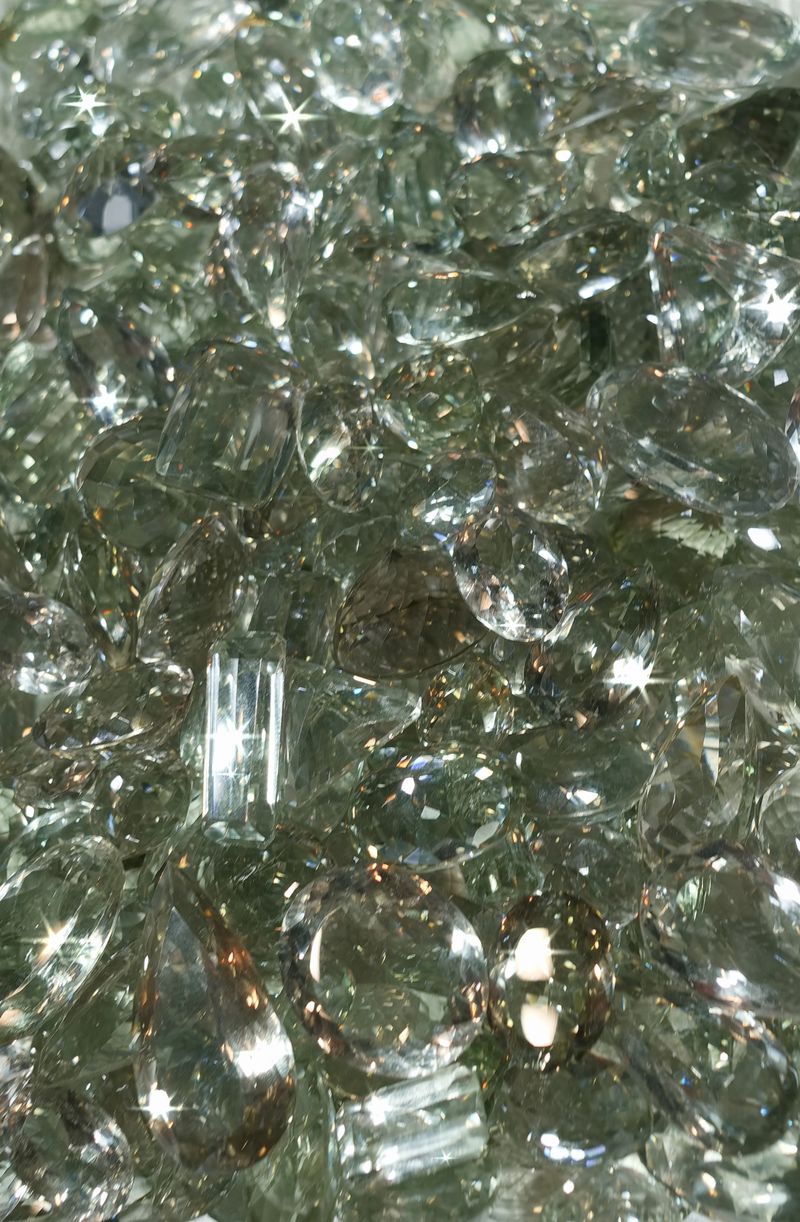 Clear gems are scattered on a flat surface, reflecting light. The scene shows many shapes and sizes, highlighting the brightness in the setting. No specific location is evident.