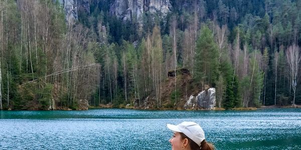 Woman in white cap near a lake surrounded by forest and cliffs.