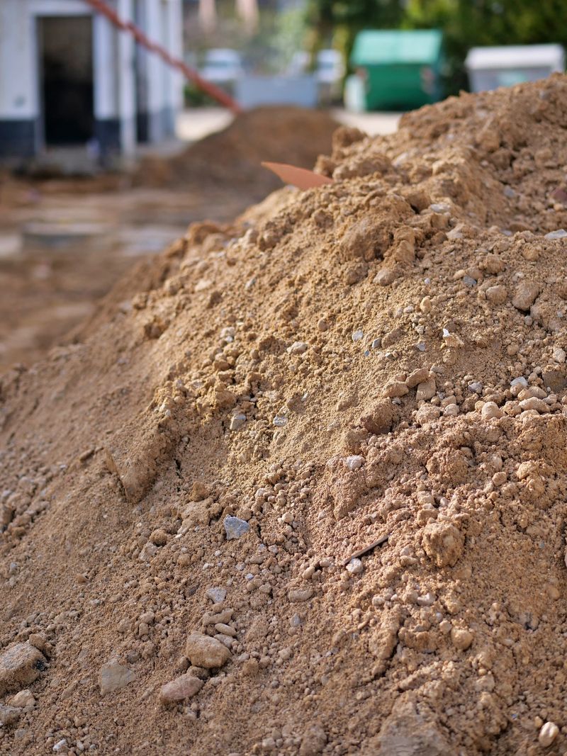 Construction site dirt pile close-up, large mounds of excavated earth and gravel, brown soil texture with small rocks, industrial site background with blurry green dumpsters, outdoor renovation work scene, heap of sand and debris, shallow depth of field earthwork, building site landscape, raw construction materials, gravel pit aesthetic, pile of dirt on pavement, urban development project.