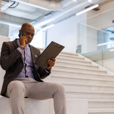 Man sitting on stairs talking on phone and holding clipboard.