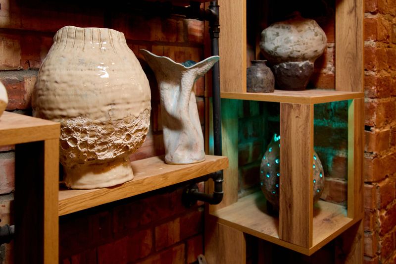 A variety of unique, textured handmade ceramic vases and art pieces displayed on wooden shelves against a rustic red brick wall in a pottery studio. The collection features different glazes, shapes, and textures, highlighted by soft indoor lighting and industrial pipes.