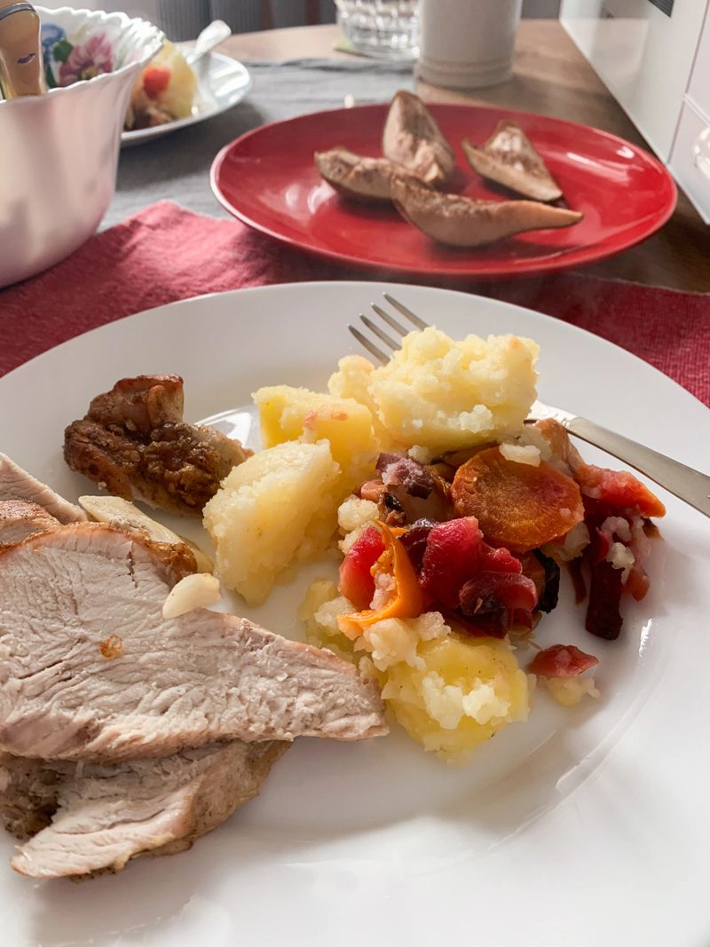 A plate holds sliced turkey meat, mashed potatoes, and a mix of colorful vegetables. In the background, baked potato wedges are served on a red plate. The setting is casual and inviting.