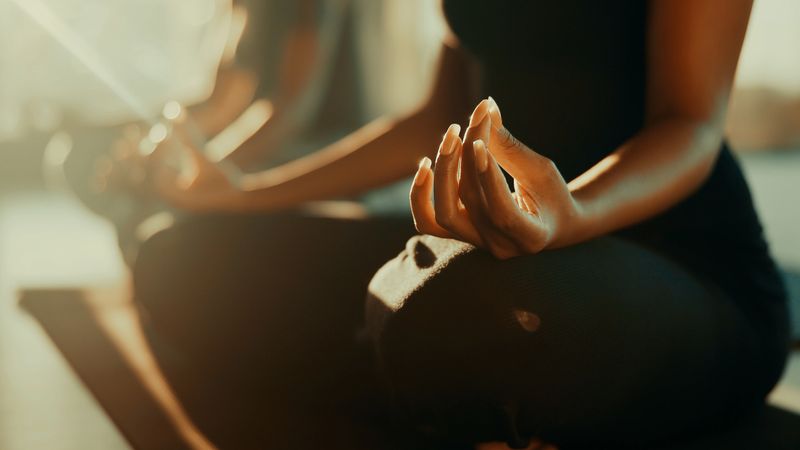 Two young women sitting cross-legged outdoors at sunset performing yoga meditation. They wear dark athletic attire, with hands in mudra gesture, radiating calm and focus in a natural environment