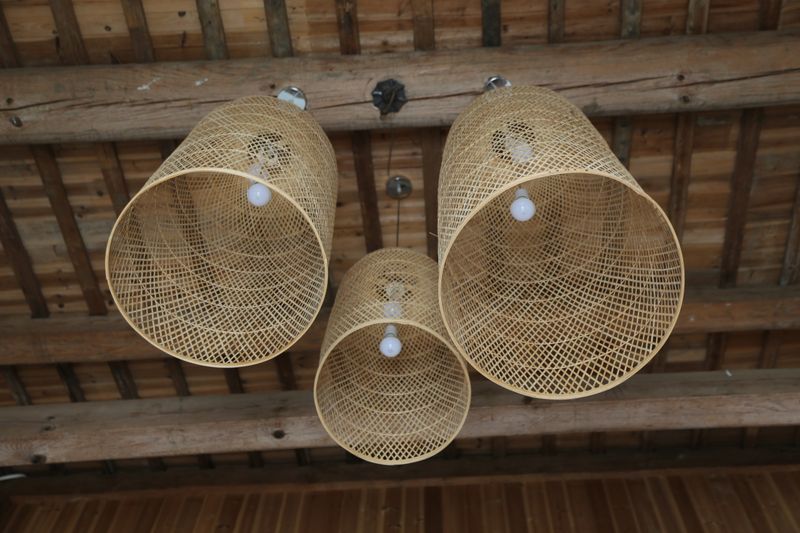 A low angle view of three beautifully crafted rattan or bamboo lampshades suspended from exposed wooden beams. The warm, natural textures of the woven material and the raw wood ceiling create a cozy, bohemian, and rustic architectural detail.
