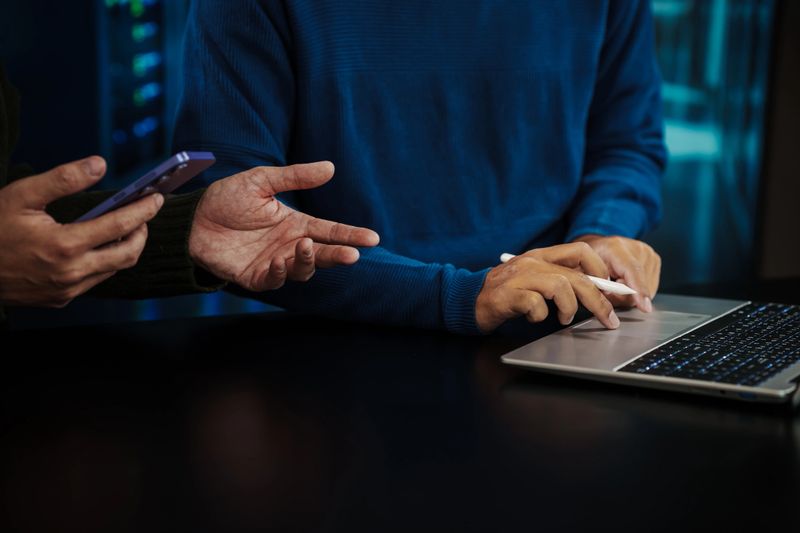 Close-up of coworkers hands at desk using laptop and smartphone in server room, discussing data or cybersecurity work with blue lighting.