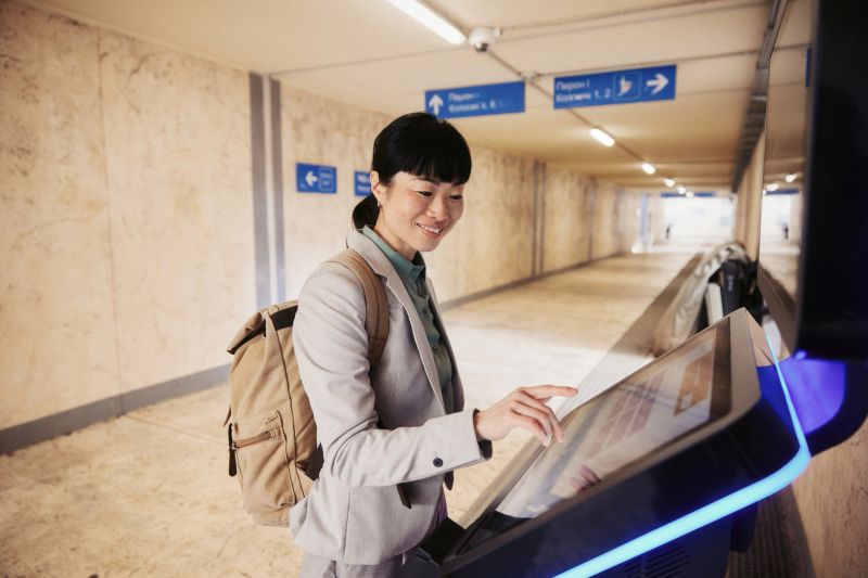 Young adult woman at a train station uses a self-service ticket kiosk while carrying a backpack; focused and confident traveler interacting with a digital touchscreen for public transport and travel planning.