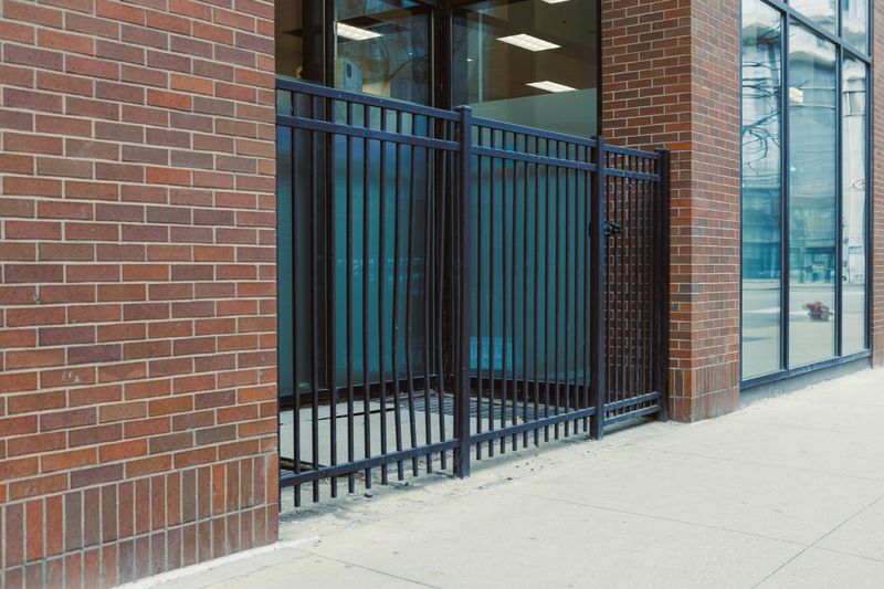 A black metal fence stands near a brick building with large glass windows. It is situated on a city street and shows parts of the sidewalk nearby. Daylight highlights the scene.