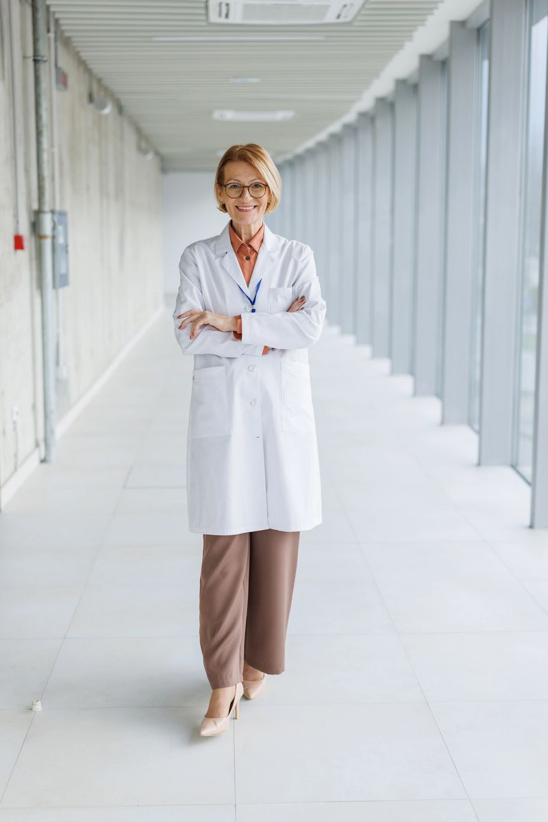 Senior adult woman in white lab coat standing with crossed arms in a brightly lit hallway