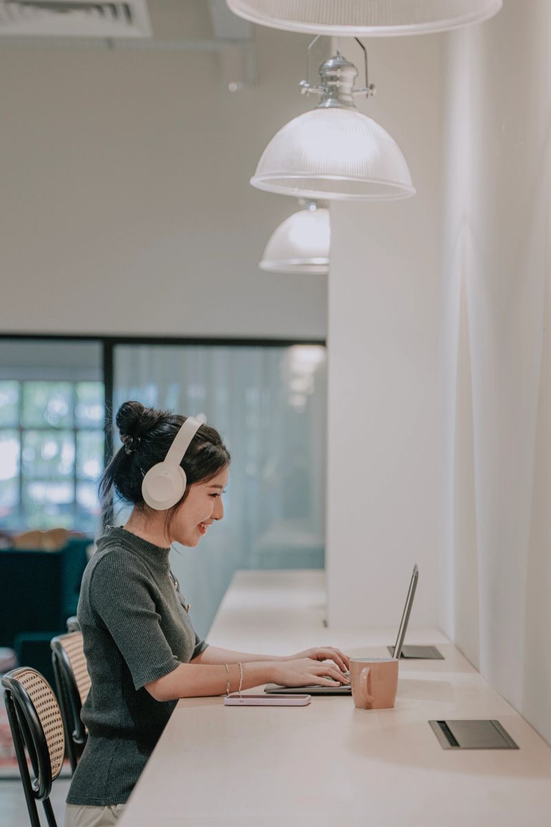 Asian woman wearing headphones typing on a laptop while working at a desk in a modern coworking workspace, representing remote work, productivity and digital communication in a contemporary work environment, Penang