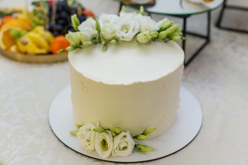 Elegant buttercream cake decorated with white flowers on festive dessert table with fruit and pastries in soft background.