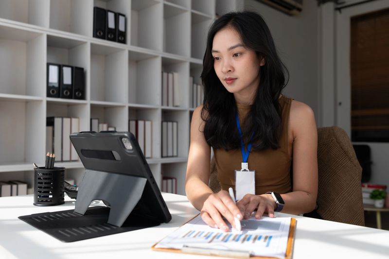 Young adult woman focusing on business documents with charts while working at an office desk with a digital tablet