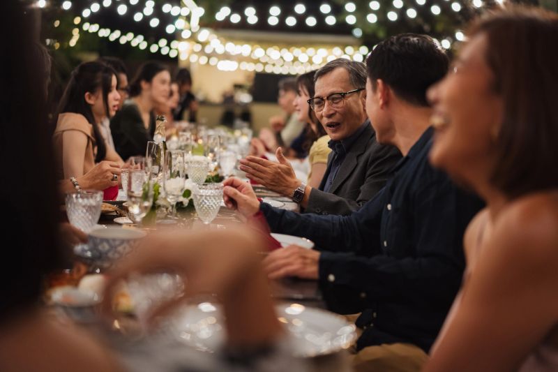 A large family enjoying dinner together at a long table in an elegant outdoor setting, sharing conversation and a meal in a warm evening atmosphere.