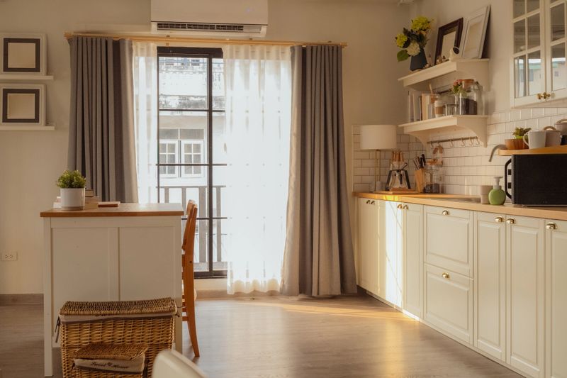 Bright modern kitchen and dining area with wooden chairs and island table in cozy apartment interior, natural sunlight through window creating warm minimalist home lifestyle and stylish domestic space.