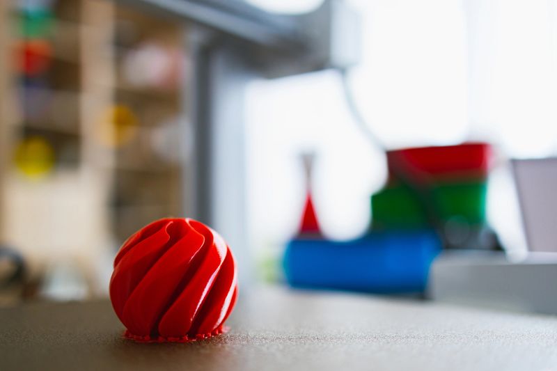 A bright red, intricately designed sphere sits on the print bed of a 3D printer, with other colorful objects blurred in the background