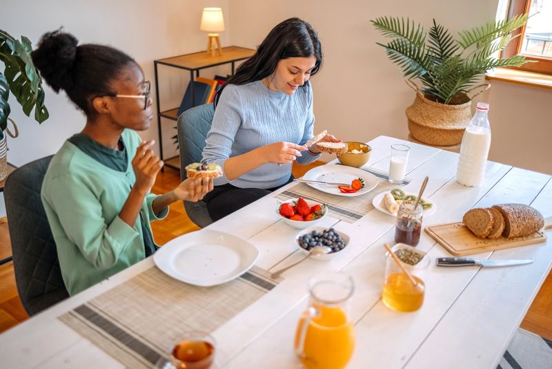 Female friends living healthy lifestyle preparing breakfast together with fruits, honey, butter and bread.