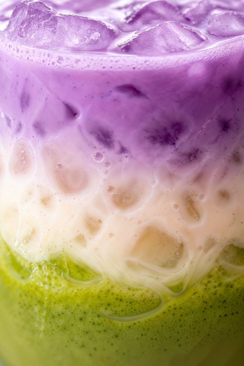 Macro close-up of a colorful ube and matcha latte with ice, featuring beautiful layered tones of purple, creamy white, and vibrant green. This visually striking beverage combines ube purple yam and matcha green tea, two ingredients widely appreciated in modern café culture for their natural color and antioxidant properties. The footage highlights the layered liquids, smooth textures, and refreshing iced appearance that make this drink both appetizing and visually engaging. Recipes combining ube and matcha are increasingly popular in plant-based cuisine, wellness-focused lifestyles, and contemporary coffee shop menus. This eye-catching drink is ideal for café promotions, beverage advertising, smoothie bars, and social media food content.