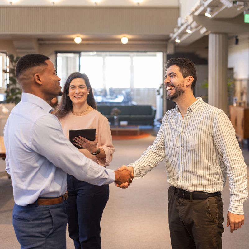 Two business colleagues shake hands warmly while a coworker smiles and holds a tablet in a bright, modern office, conveying teamwork, professional connection, networking and positive collaboration.