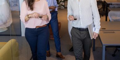 Three colleagues walking and talking in an office setting with drinks.
