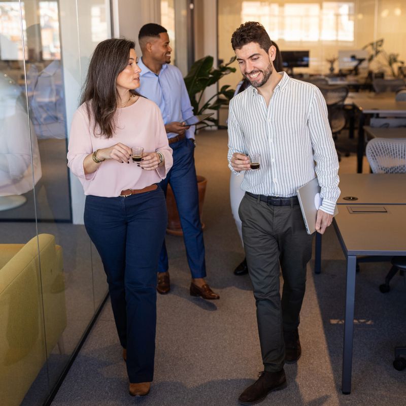 Business colleagues walk through a modern open-plan office chatting and smiling while holding coffee and a laptop, conveying teamwork, casual collaboration and a relaxed professional atmosphere.