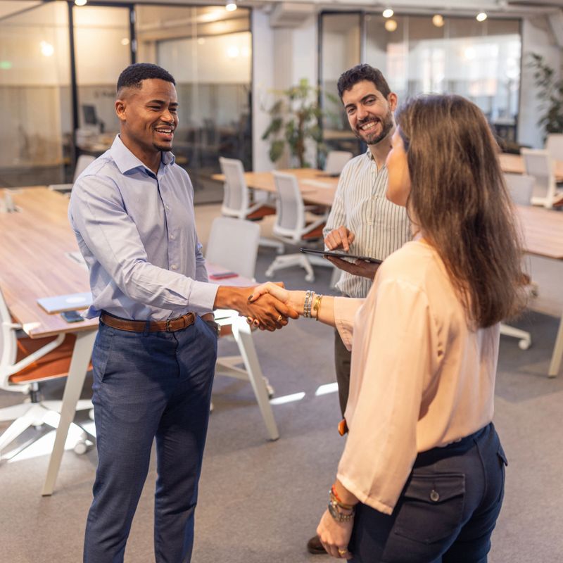 Three smiling professionals meet in a modern open-plan office, exchanging a handshake and introductions while a colleague looks on with a tablet, conveying collaboration, trust and teamwork.