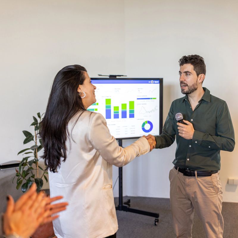 Colleagues shake hands after a business presentation in an office, with a speaker holding a microphone, charts on a screen and coworkers applauding to celebrate teamwork and successful results.