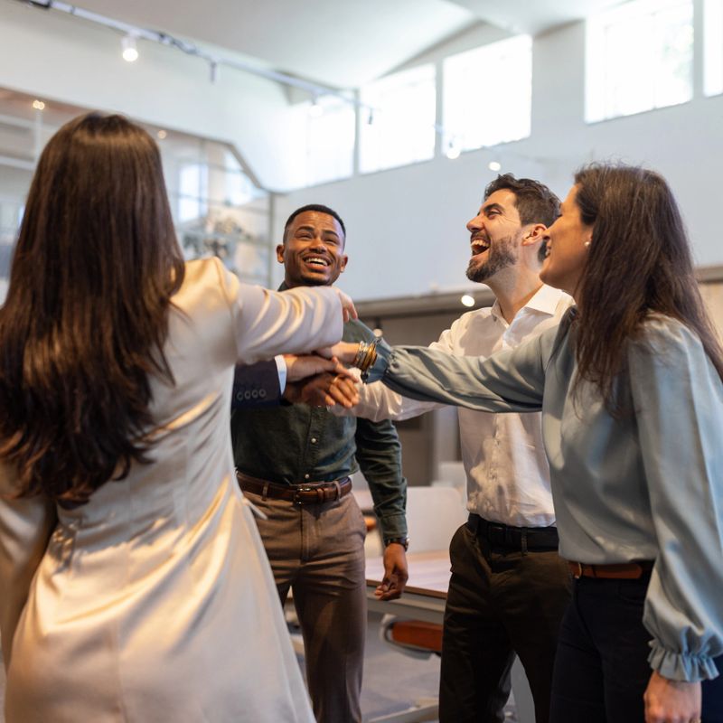 A diverse group of colleagues join hands in a team huddle, laughing and celebrating success in a bright modern office, conveying teamwork, collaboration, and positive workplace energy.