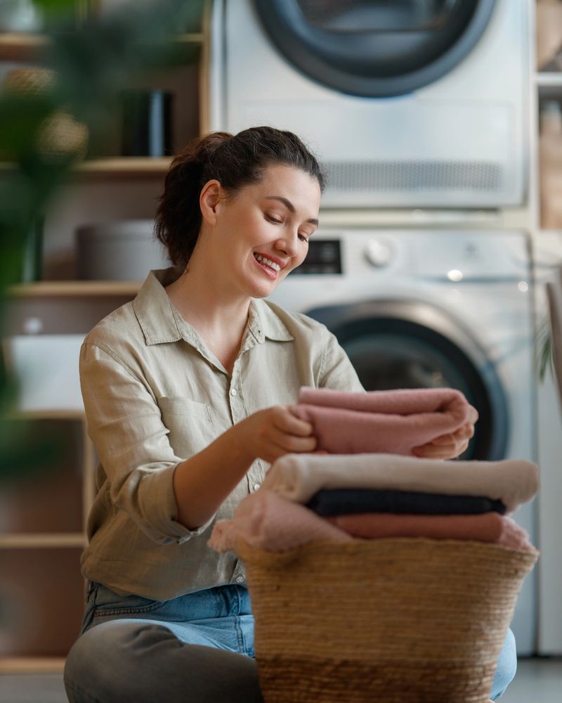 Cheerful young woman folding freshly washed clothes in a stylish laundry room, capturing a sense of domestic happiness and everyday comfort. This image evokes a feeling of satisfaction, productivity, and the warmth of a well-kept home, making it perfect for promoting household products, home care services, or lifestyle brands.