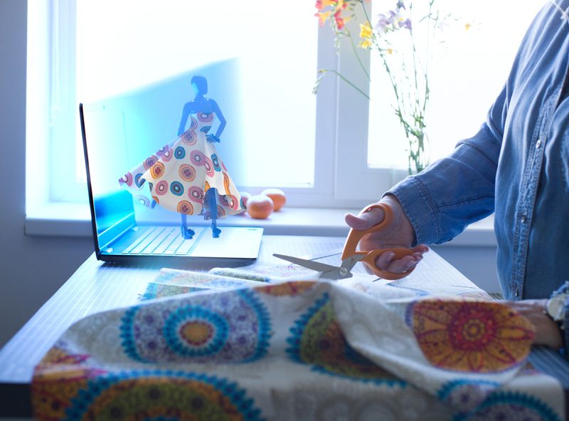 An augmented reality (AR) holographic projection emerge from a laptop to visualize a 3D dress model. The image illustrates the seamless integration of artificial intelligence in creative industries, showing how digital twin technology and smart visualization assist in the precise manual cutting of fabric patterns. High-tech meets traditional craftsmanship. Photography and 3D rendering combined.