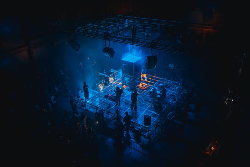 Wide high angle view of a live electronic band performing on an illuminated stage with dramatic blue lighting and audience surrounding the venue, Bangkok
