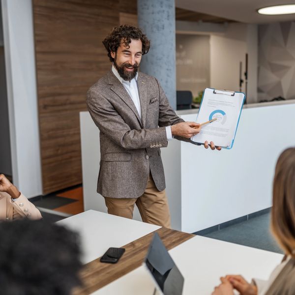 Man presenting data on a clipboard to colleagues in a meeting room.