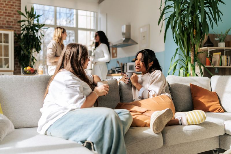 Two women sitting on a sofa and talking while other friends chat near the kitchen in a bright modern apartment. The scene captures a relaxed home gathering and casual time spent with friends.
