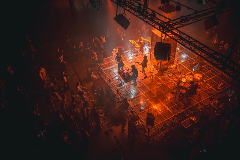 High-angle view of a live band performing on an illuminated concert stage with dramatic stage lighting, haze, instruments, and audience inside a dark indoor music venue.