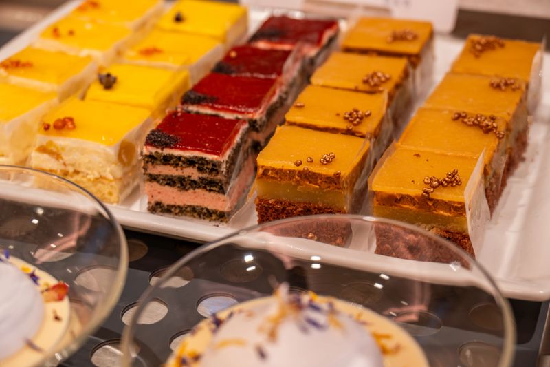 A variety of colorful dessert cake slices arranged neatly on a buffet tray. The selection includes layered sponge cakes, fruit-topped pastries, and creamy desserts with decorative finishes. The vibrant presentation highlights a rich and indulgent catering display.
