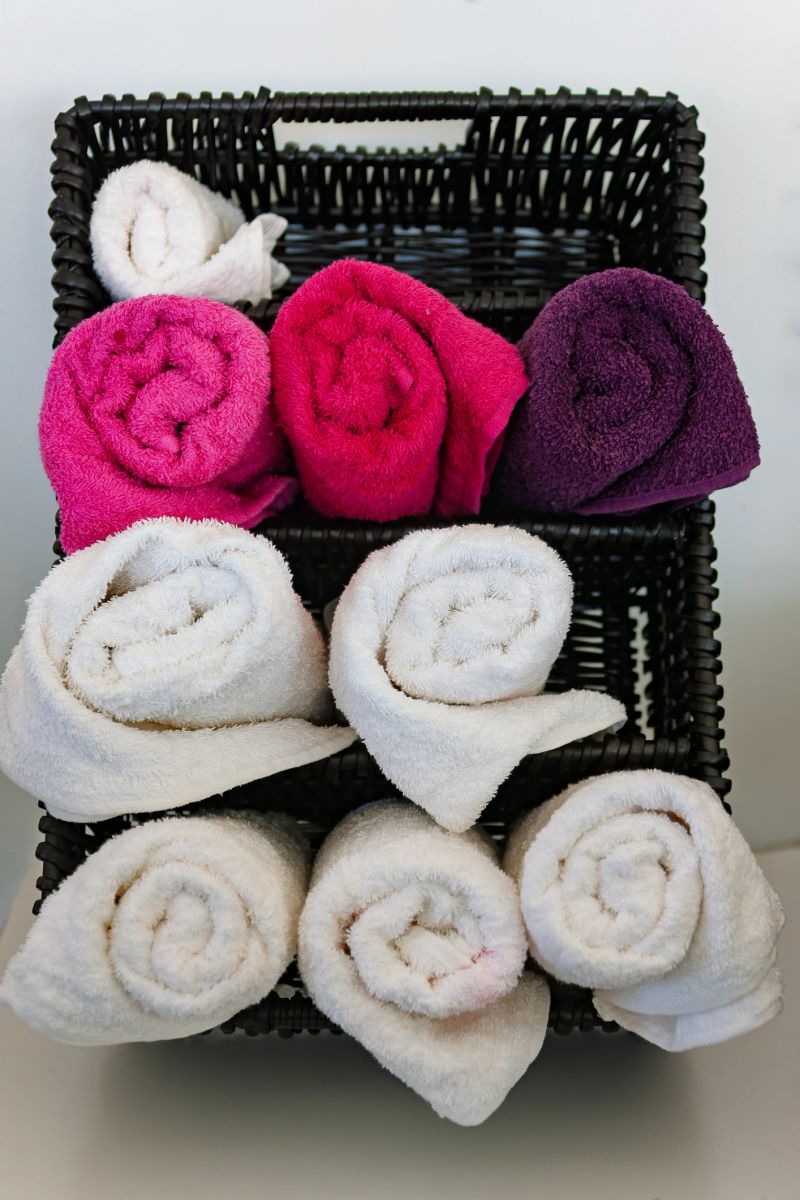 collection of white, pink, and purple cotton towels are neatly rolled and organized within the compartments of a black woven storage basket.