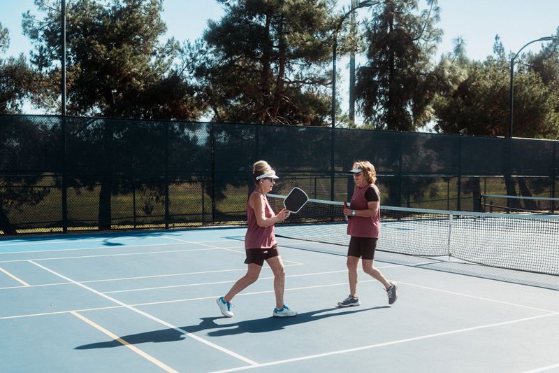 Teammates walk toward each other and talk while resetting for the next rally during a women's doubles pickleball match.