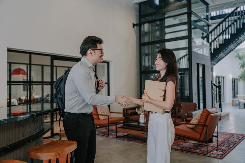 Asian businessman wearing a backpack shakes hands with an Asian woman holding a folder in a modern office lobby, suggesting a job interview, business meeting, professional greeting, or appointment arrival.