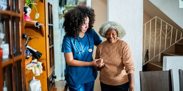 A nurse assists an elderly woman walking indoors, showing care and support.