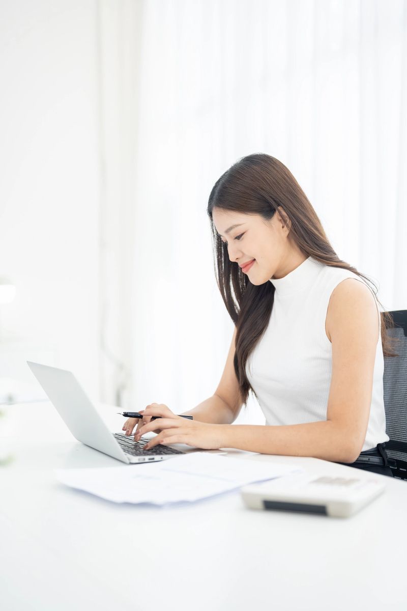 Young businesswoman working on laptop and reviewing documents in bright modern office. Online business and freelance working concept with calculator and paperwork on desk.