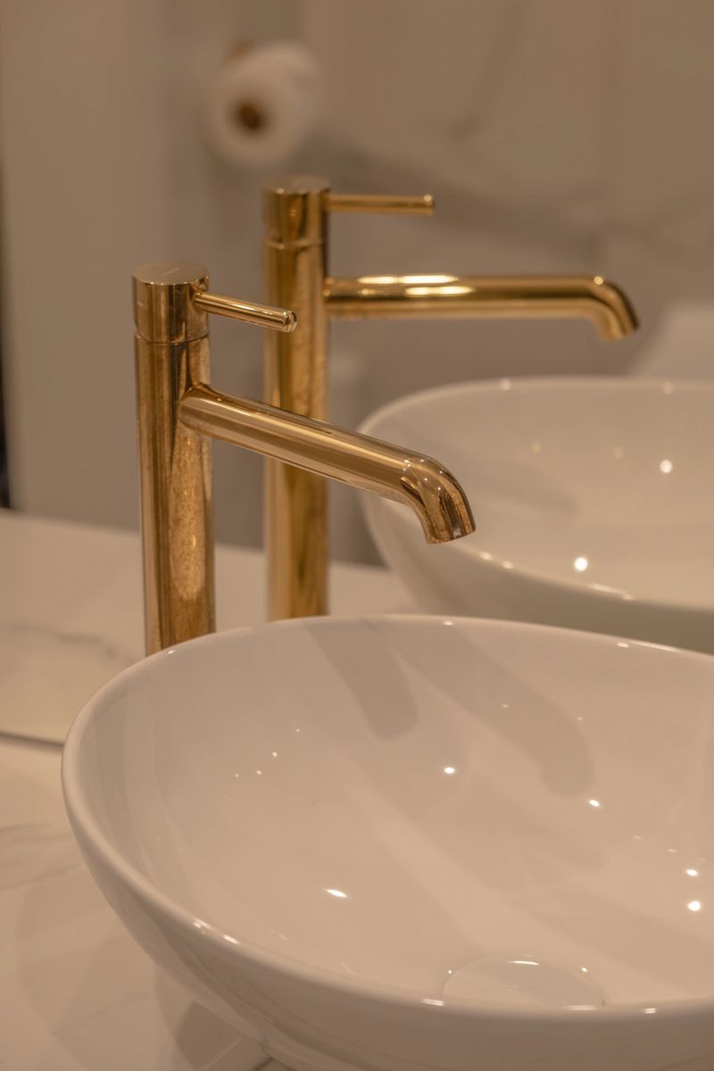 Close up of a modern luxury bathroom featuring a white ceramic vessel sink and elegant gold faucet with soft warm lighting. Clean minimal interior design with reflective surfaces creates a premium spa atmosphere. Perfect for themes of bathroom renovation, hygiene, hotel interior, real estate marketing, plumbing fixtures, and high end home decor.