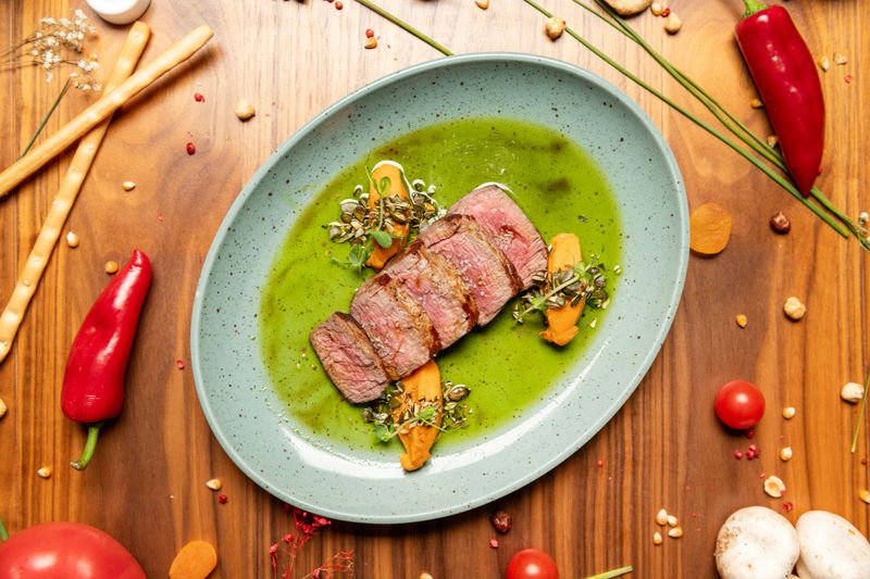 Sliced medium rare beef steak served over vibrant green herb sauce with carrot puree and seed garnish, plated on ceramic dish, rustic wooden table, top view, flat lay.