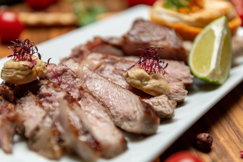 Grilled sliced lamb rack served with creamy mustard sauce and baked goat cheese on sweet potato rounds garnished with herbs and lime wedge plated on a rectangular white dish on a rustic wooden table