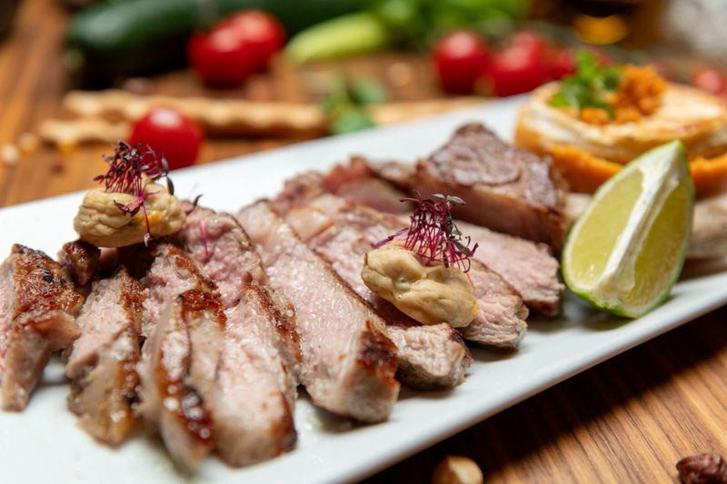 Grilled sliced lamb rack served with creamy mustard sauce and baked goat cheese on sweet potato rounds garnished with herbs and lime wedge plated on a rectangular white dish on a rustic wooden table